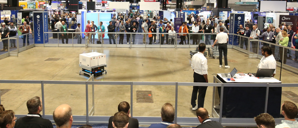 Demonstration area with autonomous mobile robots (AMRs) on display, surrounded by raised platforms with spectators and technical staff observing.