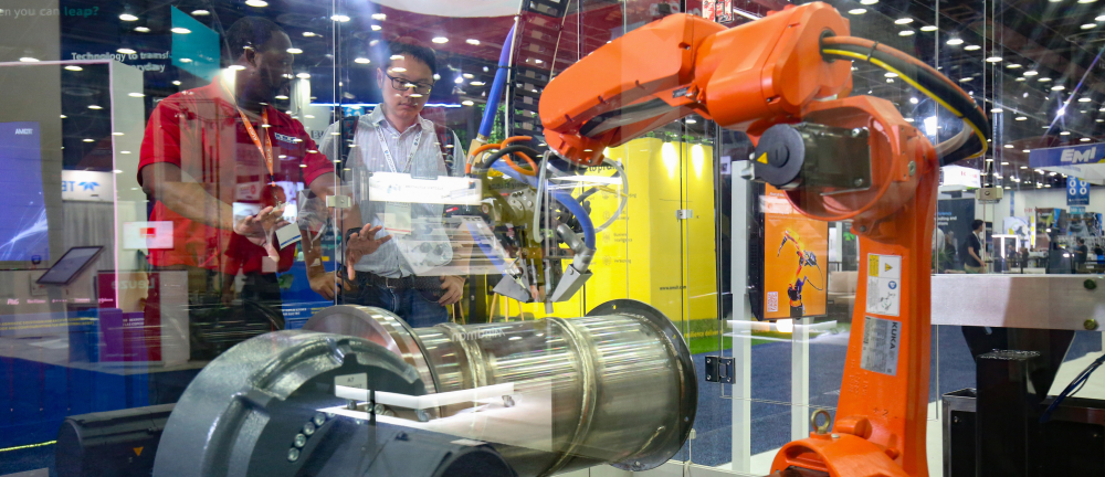 Orange industrial robotic arm performing automated welding or manufacturing task on a large metallic cylindrical component on the show floor.]