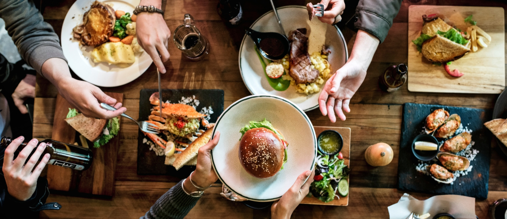 Alt text: Banner image of an overhead view of a group lunch, showing multiple people reaching for food with sandwiches, salads, and drinks on a table.