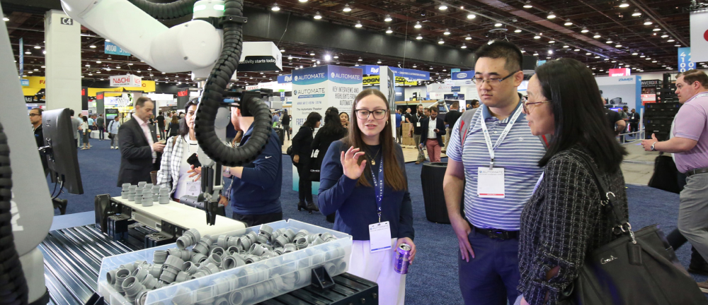 Trade show attendees observe a robotic arm demonstration handling parts at Automate
