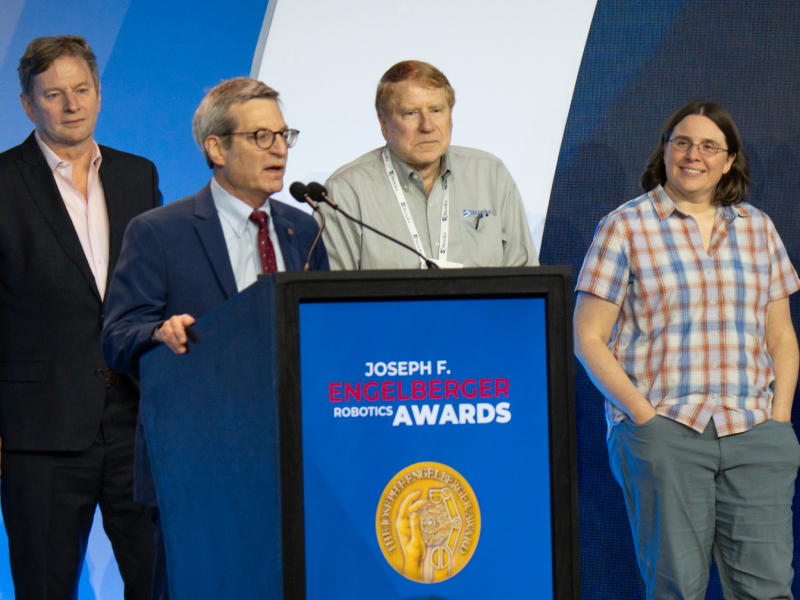 Four people stand on stage at the Joseph F. Engelberger Robotics Awards ceremony. One man speaks at a podium with the award logo, while the others stand behind him, listening and smiling.