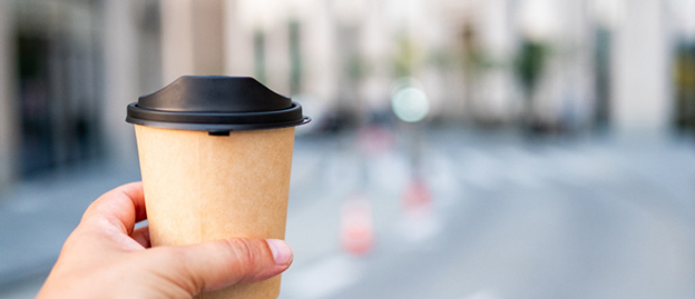 Hand holding a tan paper coffee cup with black lid against a blurred urban street background.