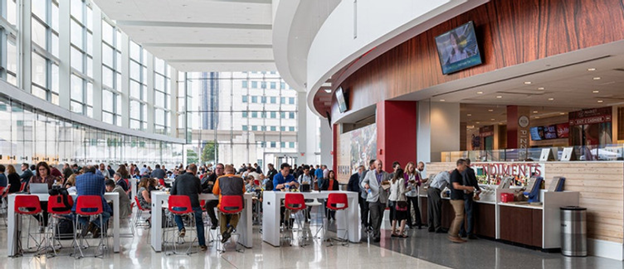 Busy food court at Huntington Place Detroit with people dining at red and white seating areas and standing in line at food vendors. Large windows provide natural light and city views.