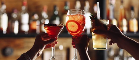 Three hands toasting with colorful cocktails at a bar and bottles blurred in the background
