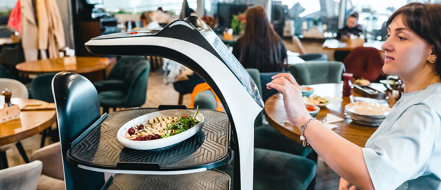 Service robot delivering a plated salad to a restaurant table while a customer reaches to interact with it. The robot has a sleek black-and-white design with a built-in serving tray.