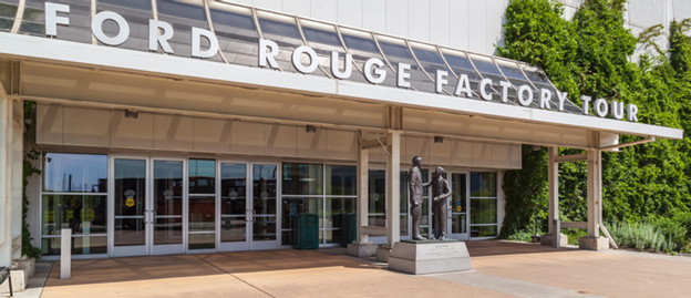 Entrance to the Ford Rouge Factory Tour featuring glass doors under a white overhang with large lettering, and a metallic sculpture of factory workers near the doorway.